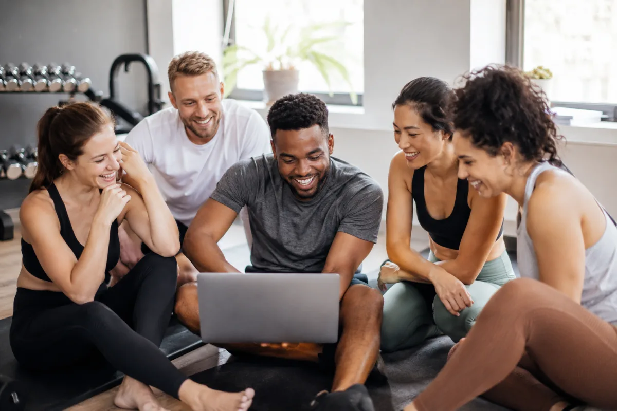 Group of personal trainers sitting on a gym floor laughing and looking at a laptop together