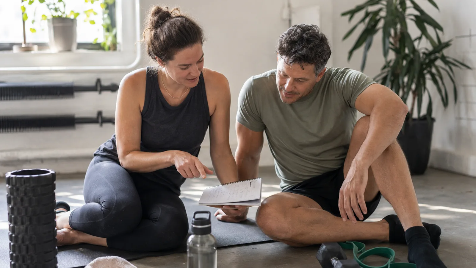 Personal trainer reviewing a training plan notebook with a client on the floor of a bright studio gym