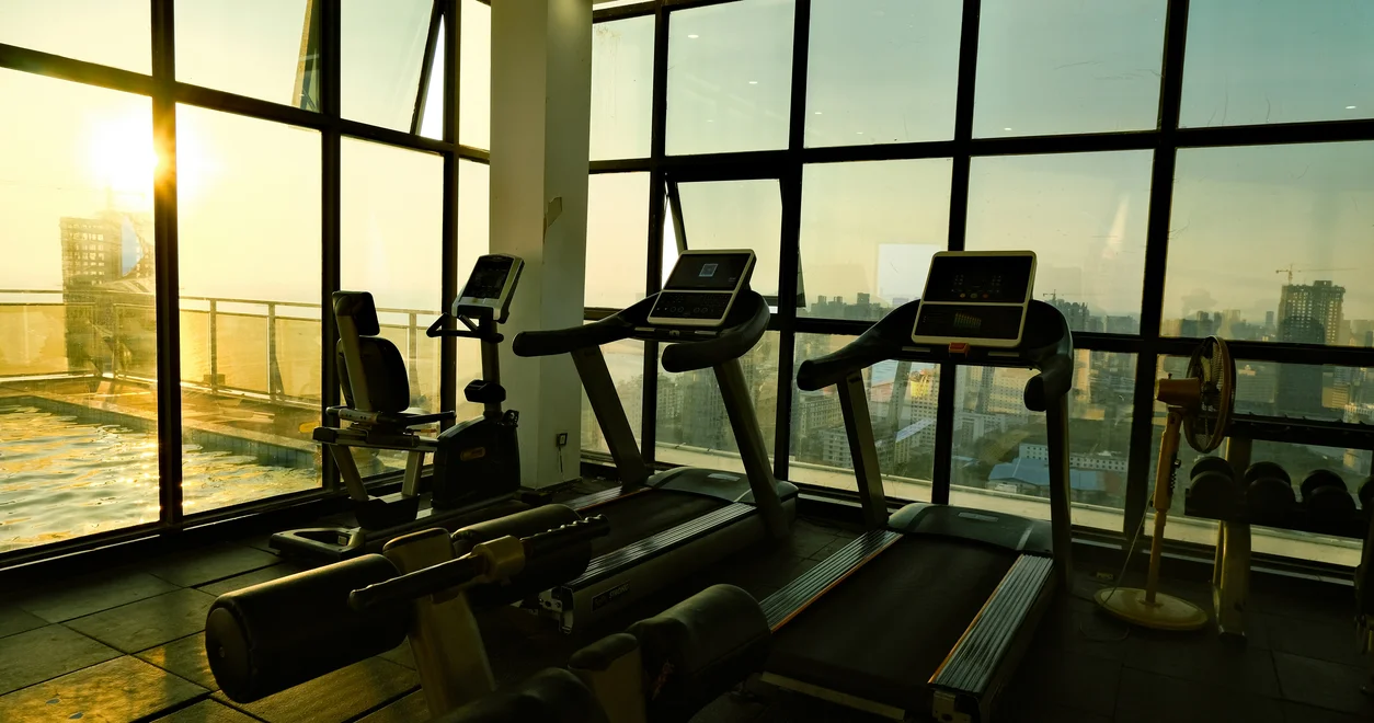 Gym at sunrise with cardio machines facing a window over a city skyline
