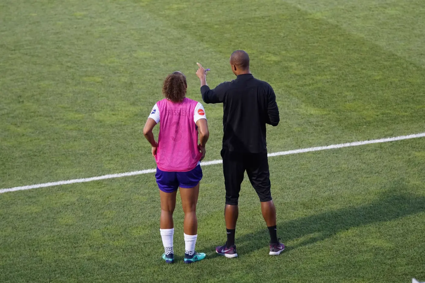 A coach standing beside a player on the pitch, pointing ahead as he gives her a short verbal cue during a session