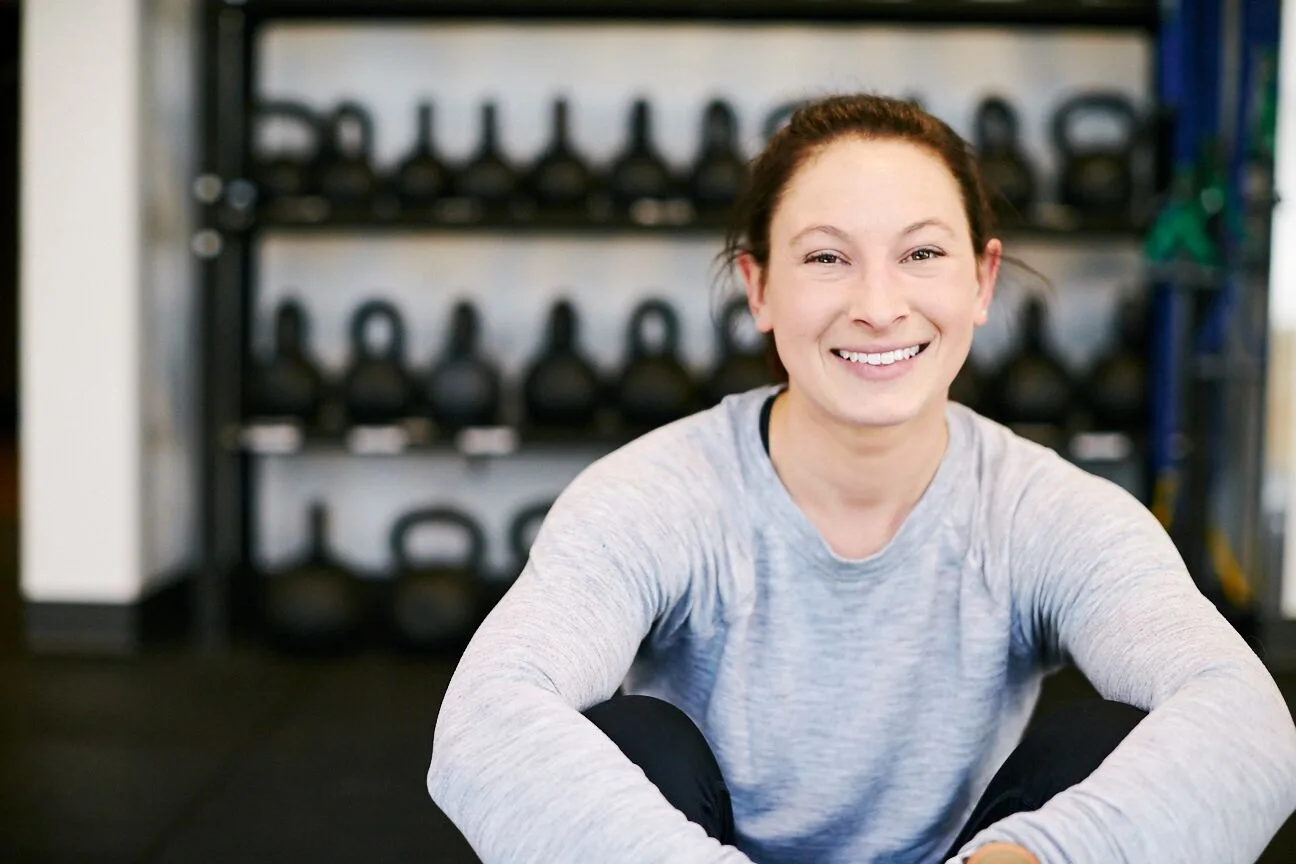 Coach Liza Duddy smiling in her Duddy Performance strength and conditioning facility, kettlebells on the rack behind her