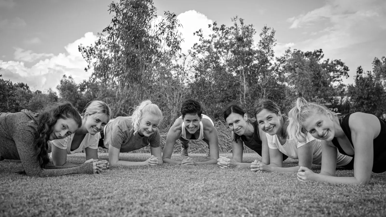 Outdoor group training session with seven smiling clients holding a plank on the grass