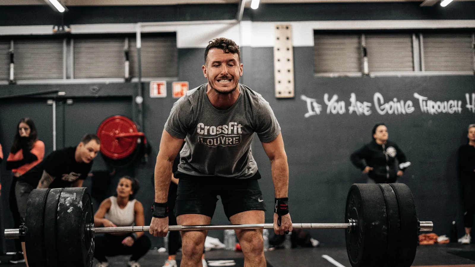 A lifter gripping a loaded barbell at the start of a deadlift while a group watches, the hip hinge that carries most of coaches' hamstring work