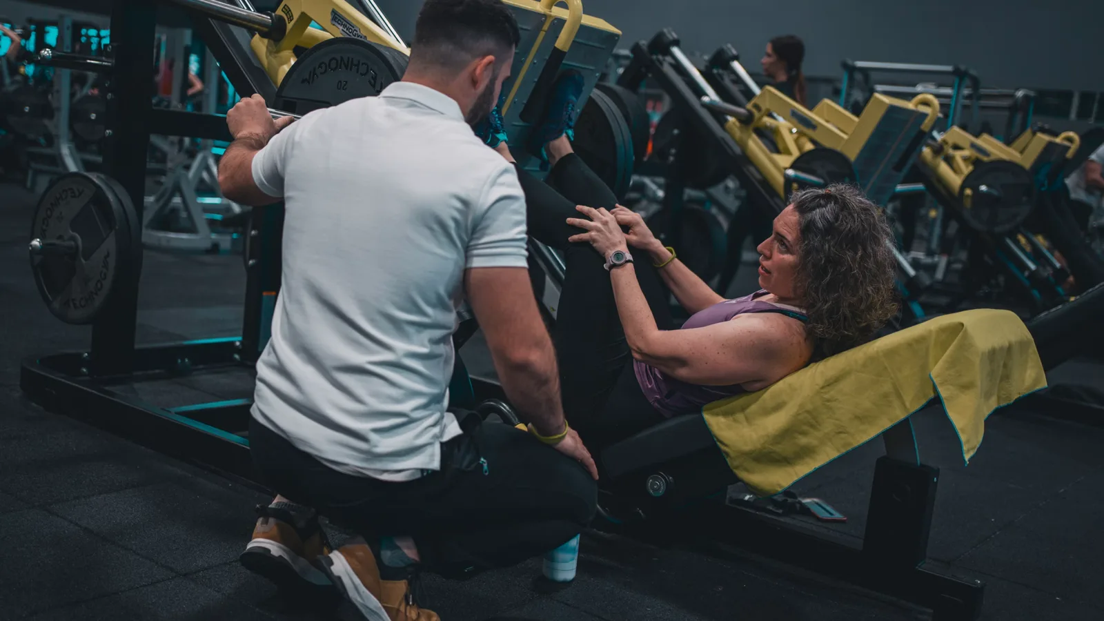 Personal trainer coaching a client through a leg press session in a busy gym