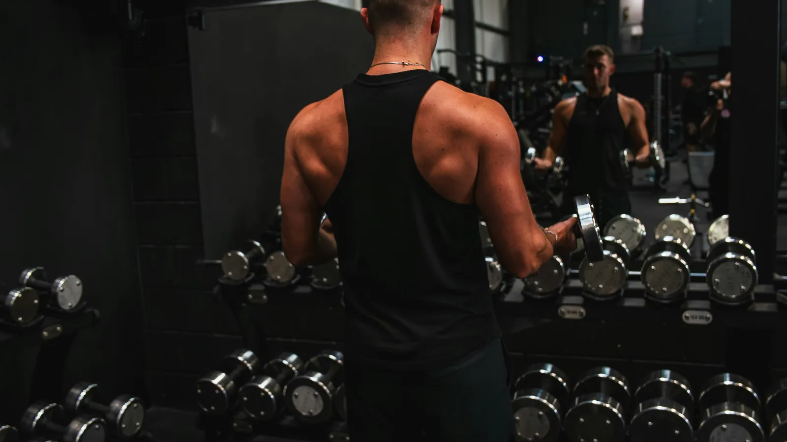 A physique athlete doing a dumbbell curl in a gym, checking his condition in the mirror behind the rack