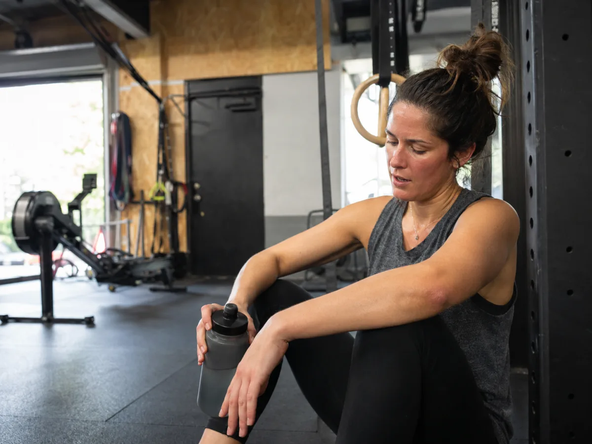 Woman in a grey tank top resting with a water bottle against a squat rack after a workout in a gym