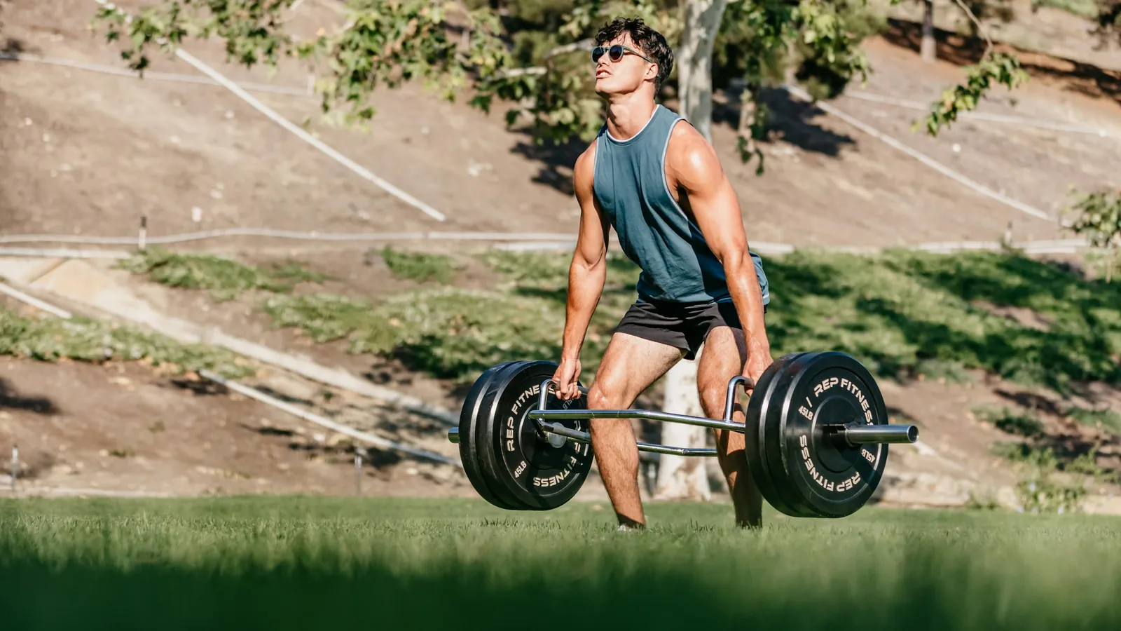 A man setting up a trap bar deadlift on the grass in a park, the movement that climbed most in coaches' programs in September 2026