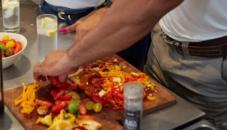 Hands slicing red and yellow capsicum on a wooden board in a kitchen, beside a bowl of cherry tomatoes and glasses of iced water