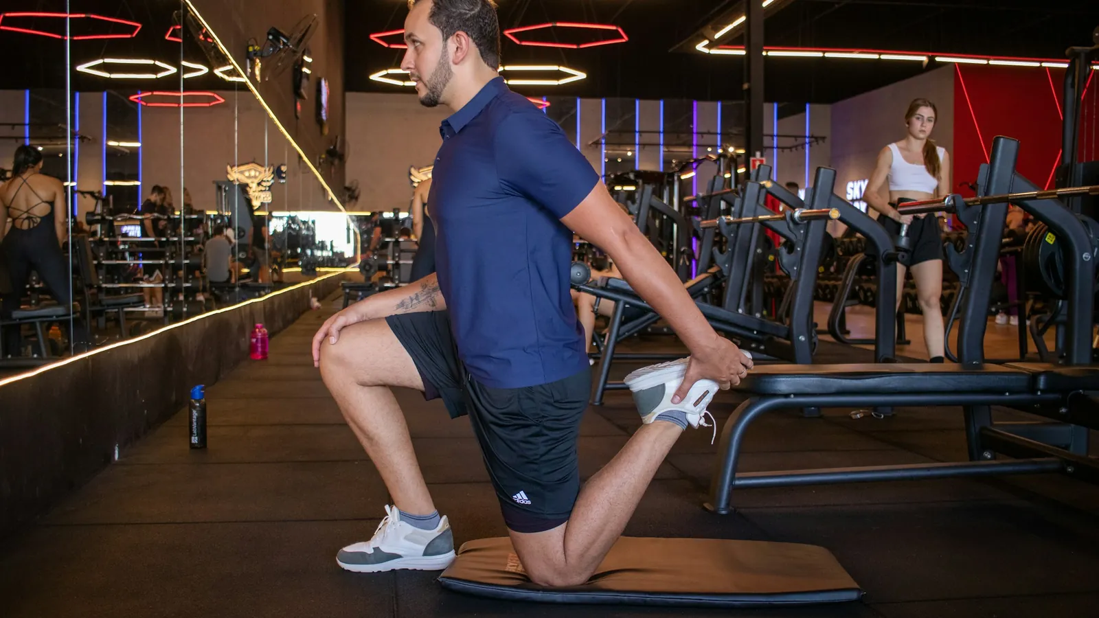 A man in a gym kneeling on a mat and holding his foot behind him in a quad stretch, the kind of warm-up drill coaches program before lifting