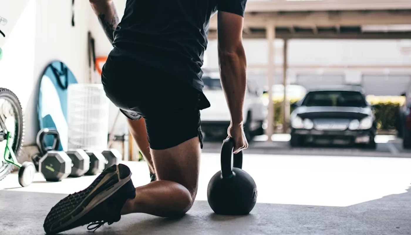 Client kneeling in a home garage gym holding a kettlebell, with a rack of dumbbells beside him, the kind of minimal equipment most online coaching programs run on
