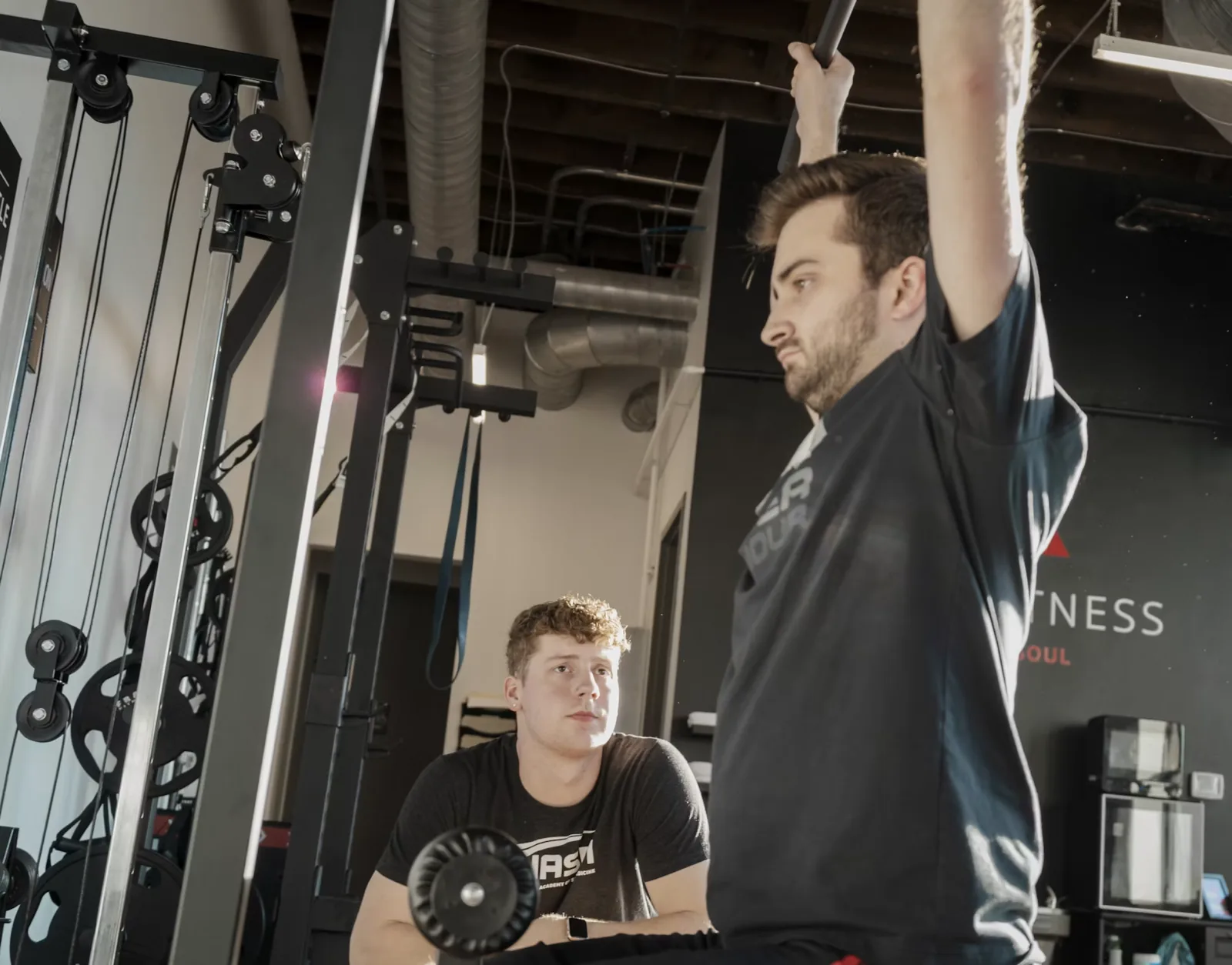 Trainer in an NASM shirt sitting back and watching a client work through a set on a cable machine in a small studio gym
